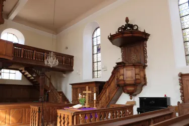 Eine alte Kirche mit Holzverzierungen und einer Empore. Im Vordergrund steht ein Altar mit einem Kreuz und farbenfrohen Blumen.
