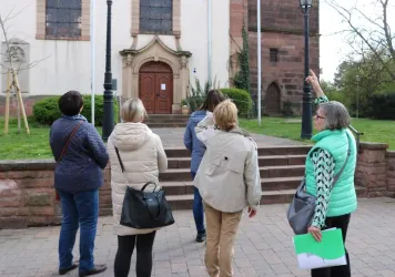 Eine Gruppe von fünf Personen steht vor einem historischen Gebäude und zeigt auf die Fassade. Sie scheinen interessiert und diskutieren darüber.