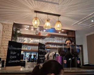 Eine gemütliche Café-Theke mit dekorativen Lampen und einem klaren Blick auf Glaswaren. Im Hintergrund steht eine Barista, die Kaffee zubereitet.