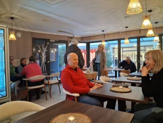 Ein gemütliches Café mit mehreren Tischen und Gästen. Die Atmosphäre ist freundlich und einladend, mit warmem Licht.