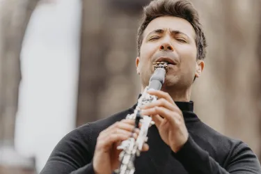 Ein Mann spielt leidenschaftlich ein Holzblasinstrument. Er trägt ein schwarzes Oberteil und konzentriert sich auf die Musik.