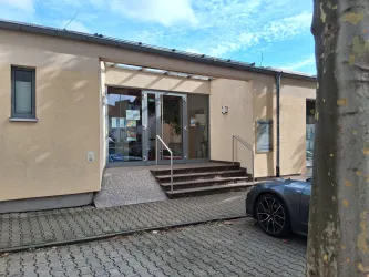 Ein modernes Gebäude mit einem klaren Eingangsbereich und Stufen. Vor dem Eingang steht ein graues Auto auf einem gepflasterten Parkplatz.