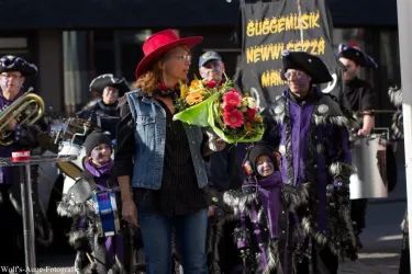 Eine Frau mit einem roten Hut hält einen Blumenstrauß. Im Hintergrund steht eine Gruppe von Menschen in Kostümen, die Musik machen.