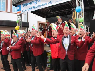 Eine Gruppe von Männern in roten Anzügen und bunten Hüten singt fröhlich auf einer Bühne. Sie wirken begeistert und nehmen an einer festlichen Veranstaltung teil.