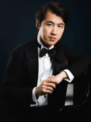 Ein junger Pianist in schwarzem Anzug sitzt neben einem Klavier. Er schaut selbstbewusst in die Kamera.