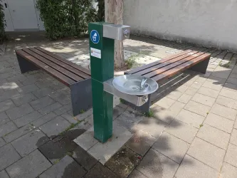 Ein Trinkbrunnen steht auf einem Pflasterweg in einem Park. Im Hintergrund sind zwei Holzbänke sichtbar.