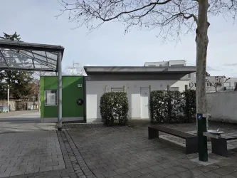 Ein moderner Sanitärbereich mit weißen Türen und einer grünen Wand. Umgeben von Büschen und einem gepflasterten Bereich.