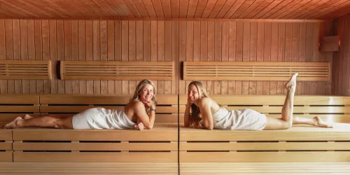 Zwei Frauen entspannen in einer Sauna auf Holzbank. Sie tragen helle Handtücher und haben Freude, während sie sich unterhalten.