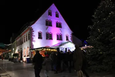 Eine festlich beleuchtete Gebäude mit bunten Lichtern und vielen Menschen vor dem Eingang. Rechts steht ein geschmückter Weihnachtsbaum.
