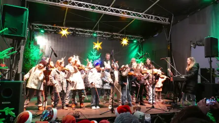 Eine Gruppe von Kindern und Erwachsenen spielt auf der Bühne Weihnachtsmusik. Die Szene ist festlich mit bunten Lichtern und Sternen dekoriert.