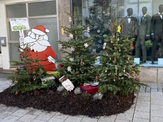 Eine festliche Weihnachtsdekoration mit Tannenbäumen und einem großen Weihnachtsmann aus Pappe. Im Hintergrund sind Schaufensterpuppen in eleganter Kleidung zu sehen.