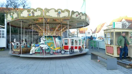 Ein nostalgisches Karussell mit bunten Fahrgeschäften steht auf einem Platz. Daneben steht eine Person in warmer Kleidung.