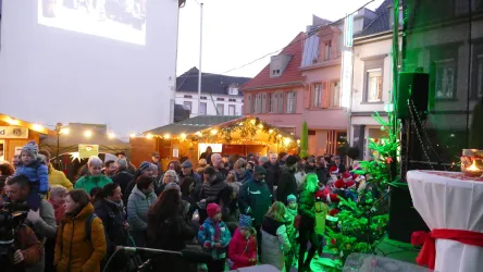 Eine große Menschenmenge versammelt sich auf einem Weihnachtsmarkt. Im Hintergrund sind festlich dekorierte Stände und beleuchtete Gebäude zu sehen.