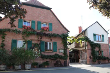 Ein charmantes, zweigeschossiges Gebäude mit roter Fassade und grünen Fensterläden. Die Fassade ist mit Pflanzen und Blumen geschmückt.
