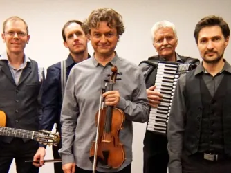 Eine Gruppe von fünf Musikern steht zusammen. Sie halten verschiedene Instrumente, darunter eine Geige und ein Akkordeon.