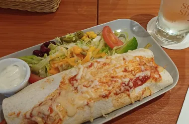 Ein großer Burrito mit geschmolzenem Käse und Tomatensauce. Dazu gibt es einen bunten Salat und einen Becher saurer Sahne.