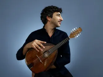 Ein Mann mit lockigem Haar sitzt und hält eine Mandoline. Er lächelt und trägt ein schwarzes Hemd.