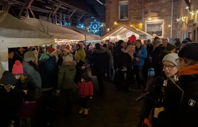 Eine belebte Marktveranstaltung bei Nacht mit vielen Menschen in warmer Kleidung. Stände mit Lichtern und festlicher Atmosphäre im Hintergrund.