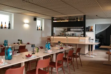 Ein modernes Restaurant mit roten Stühlen und hellen Tischen. Die Bar im Hintergrund ist dekoriert und es gibt Gläser und Flaschen auf dem Regal.