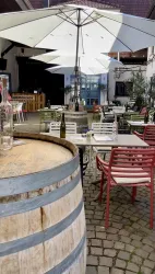 Ein gemütlicher Außenbereich eines Restaurants mit bunten Stühlen und großen Sonnenschirmen. Tische sind um ein Holzfass arrangiert, umgeben von einer einladenden Atmosphäre.