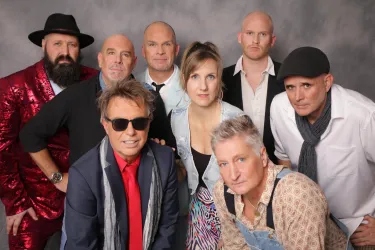 Eine Gruppe von neun Personen posiert gemeinsam für ein Foto. Die Menschen tragen verschiedene stilvolle Outfits und lächeln.