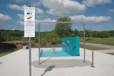 Ein moderner Wasserfilter mit blauer Wand, umgeben von einer grünen Landschaft. Die Anlage steht auf einem hellen Platz unter einem klaren, blauen Himmel.