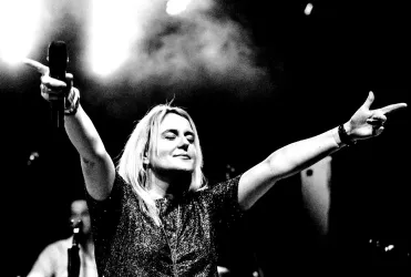 Eine Sängerin steht auf der Bühne und bringt das Publikum in Stimmung. Sie hat die Arme ausgebreitet und wirkt voller Energie.