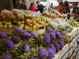Ein lebhafter Markt mit frischen Äpfeln und bunten Artischocken. Menschen stehen im Hintergrund und genießen das bunte Angebot.