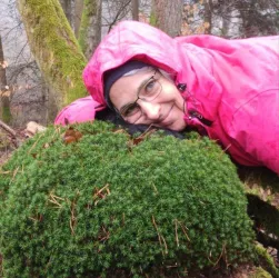 Eine Person liegt auf einem moosbedeckten Baumstamm im Wald. Sie trägt eine pinke Jacke und lächelt in die Kamera.