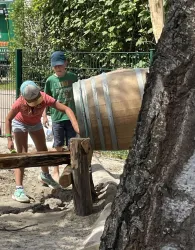 Zwei Kinder spielen im Freien mit einem großen Holzfass. Sie sind in einer Sandlandschaft und scheinen viel Spaß zu haben.