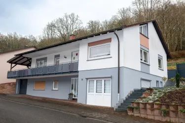 Ein modernes Haus mit einem Balkon und einer hellen Fassade. Im Hintergrund sind Bäume sichtbar, die eine ruhige Umgebung schaffen.