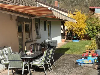 Eine gemütliche Terrasse mit einem Tisch und Stühlen, umgeben von einem grünen Garten. Im Hintergrund blüht ein gelber Strauch und es stehen Spielzeuge für Kinder bereit.