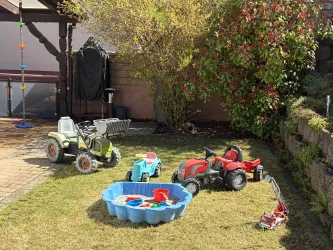 Ein Garten mit mehreren Spielzeugen, darunter kleine Traktoren und ein Planschbecken. Im Hintergrund sind Bäume und ein Pavillon zu sehen.