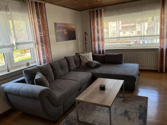 Ein gemütliches Wohnzimmer mit einem grauen Sofa und einem Holztisch. Große Fenster lassen viel Licht herein und bieten einen Blick nach draußen.
