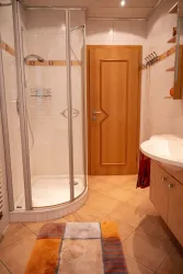 Ein modernes Badezimmer mit einer Dusche, einem Waschbecken und einer Holztür. Der Boden ist gefliest und ein bunter Teppich liegt vor der Dusche.