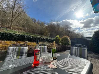 Ein gemütlicher Tisch im Freien mit Getränken und Gläsern. Im Hintergrund sind Bäume und ein klarer Himmel zu sehen.