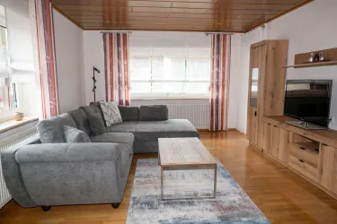 Ein gemütliches Wohnzimmer mit einem grauen Sofa und einem Holztisch. Große Fenster mit Vorhängen lassen viel Licht hinein.