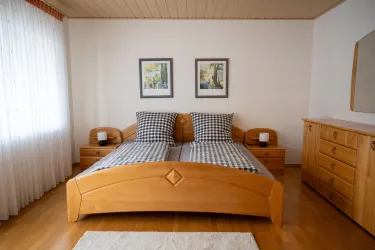 Ein helles Schlafzimmer mit einem hölzernen Doppelbett und karierten Kissen. An der Wand hängen zwei Bilder, während ein Kommode und Nachttische den Raum ergänzen.