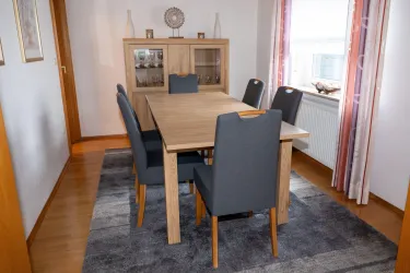 Ein moderner Essbereich mit einem holzfarbenen Tisch und sechs grauen Stühlen. Der Raum wirkt hell und einladend dank der Fenster und des Teppichs.