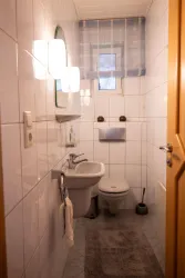 Ein kleines, helles Badezimmer mit weißen Fliesen und einem modernen Waschbecken. Das WC und ein Handtuchhalter sind gut platziert, und es gibt ein Fenster mit einem hellen Vorhang.