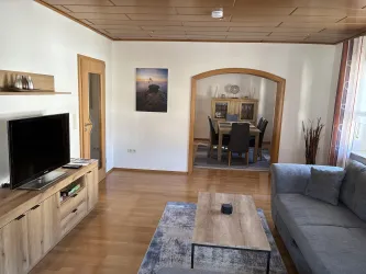 Ein helles Wohnzimmer mit einem Sofa, einem Couchtisch und einem Fernseher. Im Hintergrund ist ein Essbereich mit einem Tisch und Stühlen zu sehen.