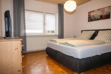 Ein gemütliches Schlafzimmer mit einem großen Bett und karierten Kissen. Ein Fernseher und ein Holzschrank vervollständigen die Einrichtung.