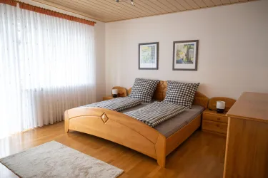 Ein einladendes Schlafzimmer mit einem Holzbett und karierten Kopfkissen. Große Fenster lassen viel Licht herein.