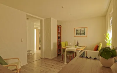 Ein modernes Wohnzimmer mit einem hellen Esstisch und bunten Stühlen. Die Wände sind in einem warmen Farbton gehalten, und es gibt einige Pflanzen zur Dekoration.