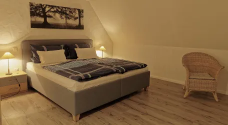 Ein gemütliches Schlafzimmer mit einem großen Bett und zwei Nachttischen. Ein Korbstuhl und ein dekoratives Bild an der Wand runden den Raum ab.