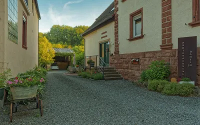 Eine ruhige Einfahrt mit einer gepflasterten Oberfläche und begrünten Bereichen. Auf der rechten Seite steht ein charmantes Gebäude mit einer Treppe und blühenden Pflanzen.