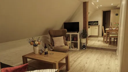 Ein gemütliches Zimmer mit Holzboden und warmem Licht. Es gibt einen bequemen Sessel, einen Couchtisch und einen Essbereich im Hintergrund.