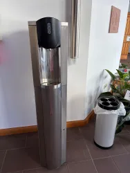 Ein moderner Wasserspender aus Edelstahl steht an einer Wand. Im Hintergrund befindet sich ein Abfalleimer und einige Pflanzen.