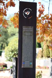 Ein steinerner Monolith mit einer Plakette, auf der "Rätselhafte Zeitzeichen" steht. Im Hintergrund sind farbige Blätter und Gräber sichtbar.