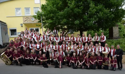 Eine große Musikgruppe steht vor einem gelben Gebäude. Die Mitglieder tragen einheitliche Kleidung in Weinrot und Weiß und halten verschiedene Musikinstrumente.